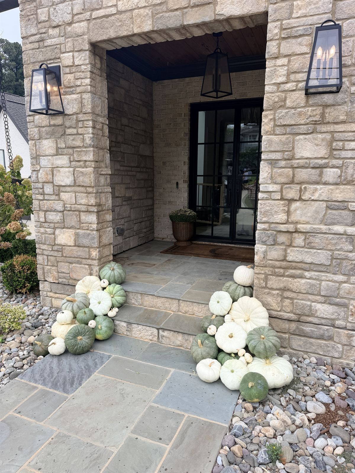 Stone entry with a black steel door and lanterns, flanked by sage and white heirloom pumpkins on bluestone steps, Wake County NC